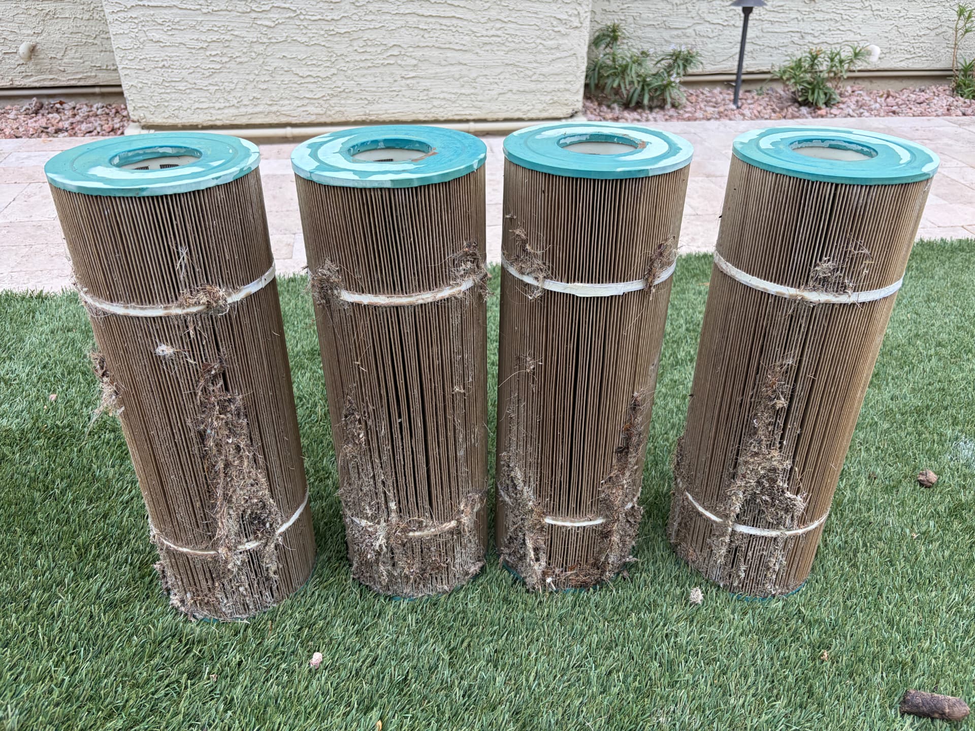 Four heavily soiled brown pool cartridge filters standing in a row on artificial grass
