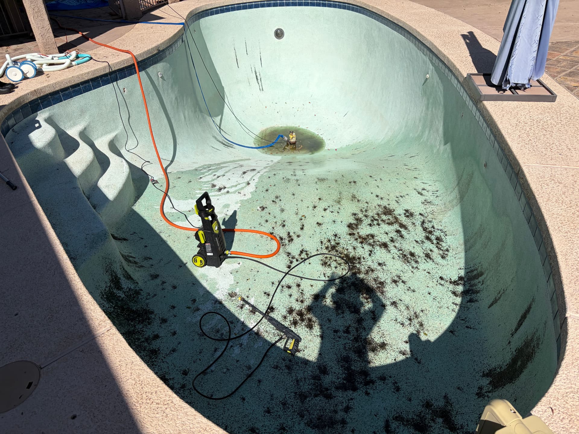An empty pool with dark algae and debris staining the plaster floor, a pressure washer standing in the shallow end